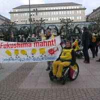 Banner Fukushima Mahnt und Demonstrierende, eine Person mit Rollstuhl und antiatom Fahne sitzt rechs vom Banner